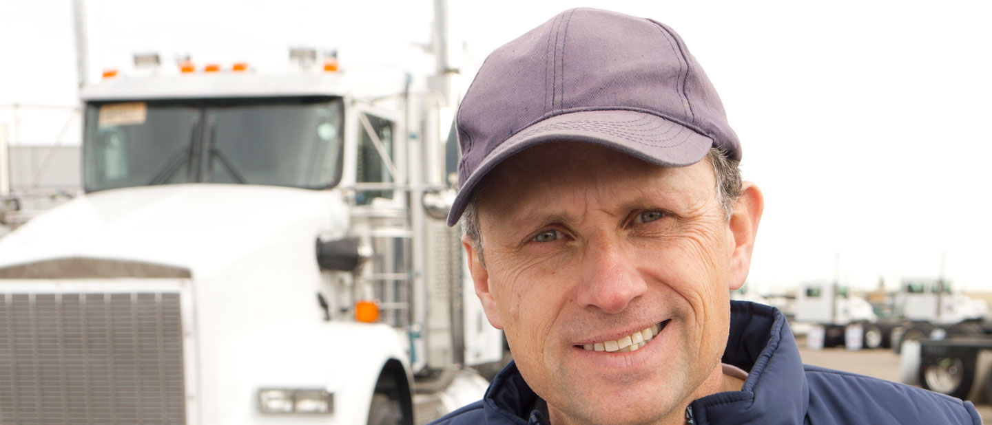 Male truck driver smiling and holding a pen and clipboard in front of his semitruck.