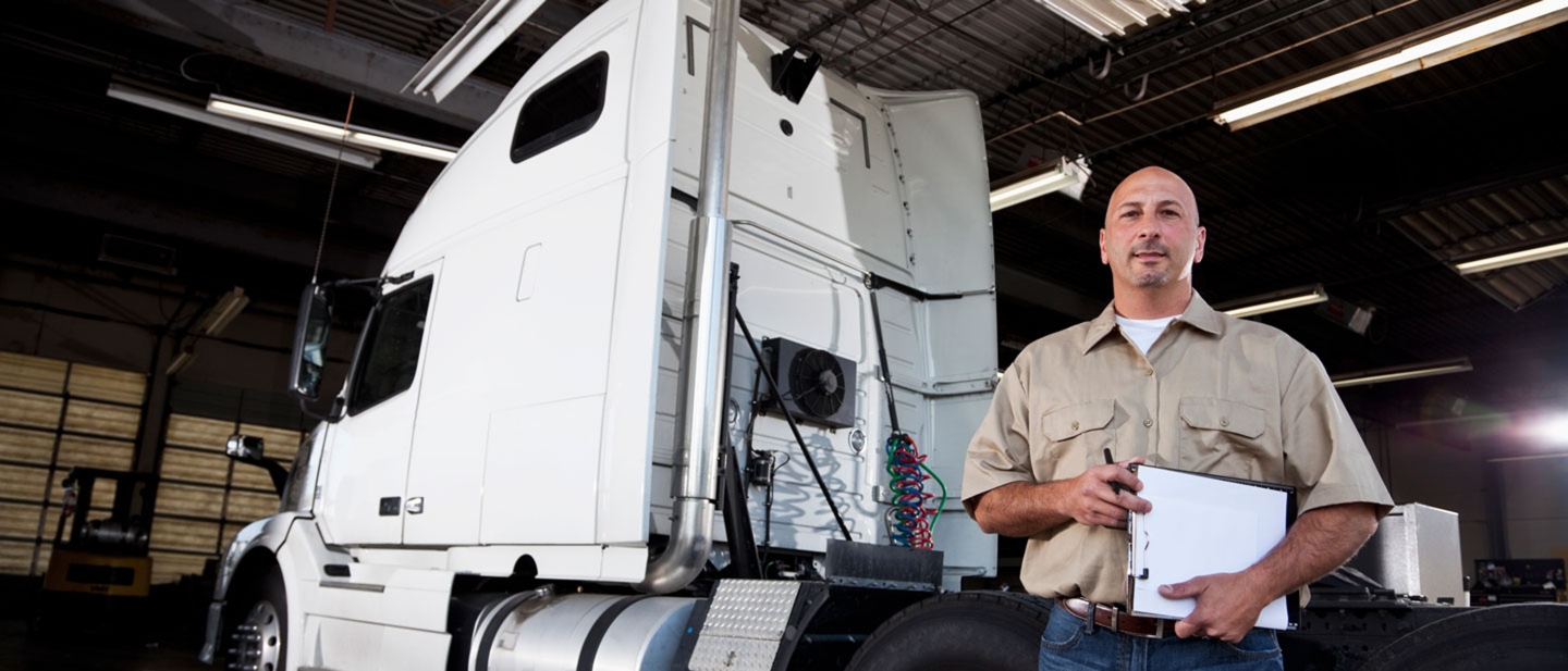 Male truck mechanic holding a clipboard stands next to a white semitruck.