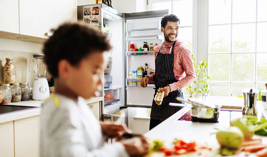 Man in background next to an open fridge while a child in the foreground is playing with food on a table.