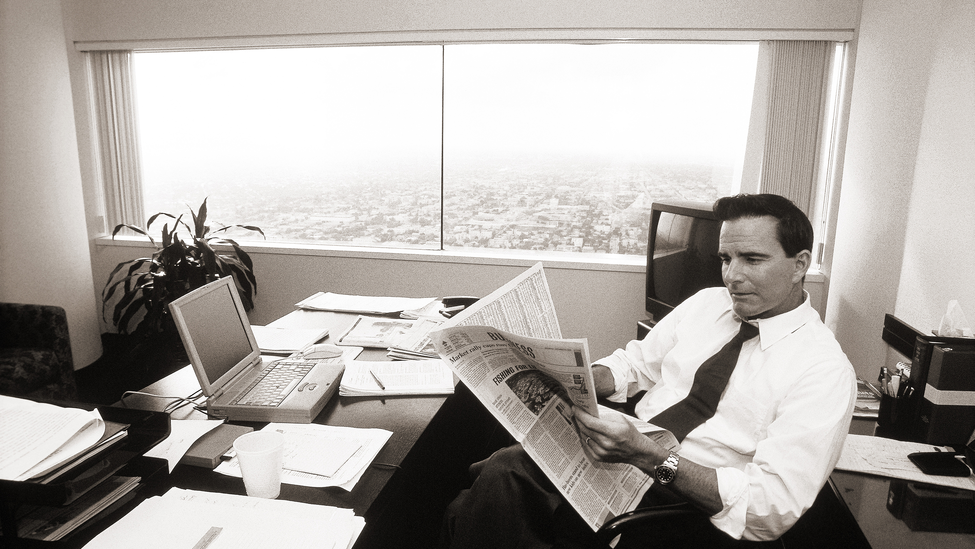 A man reads the newspaper in front of computer while sitting in a high-rise office. 