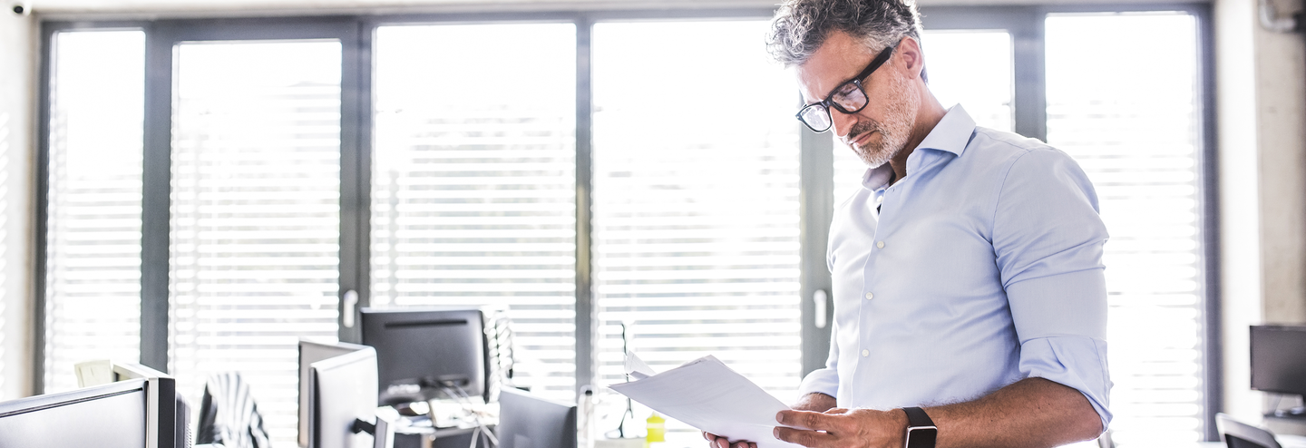 A businessman stands at a desk whilst reading some paperwork.