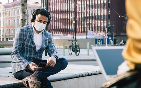 Man sitting on bench in city with a mask on and looking at his phone.