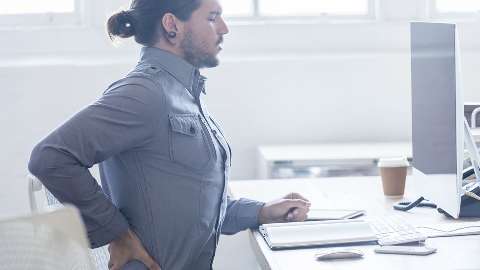 Young business man with holding his back in pain. He is sitting at a desk with a computer a keyboard and a notebook.