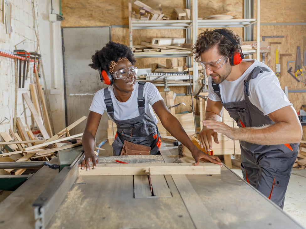 Two woodworkers wearing ear and eye protective gear use a sliding saw table to cut a section of wood.