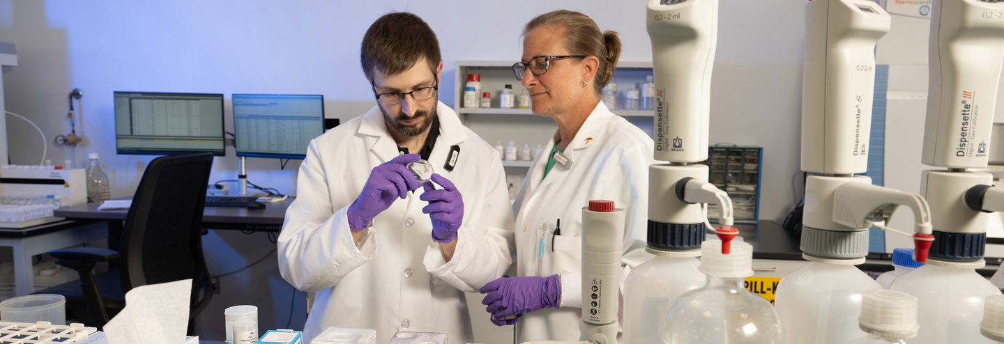 Two industrial hygienists wearing white lab coats and purple gloves looking at samples in a lab.