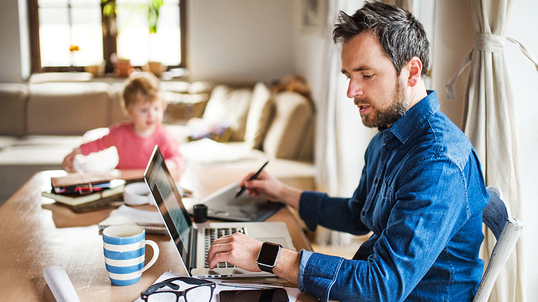 Man working from home with laptop at kitchen table with child also at the table.