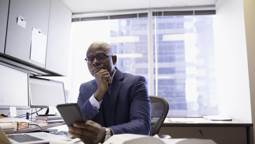 A professional man sits at his office desk looking at his phone.