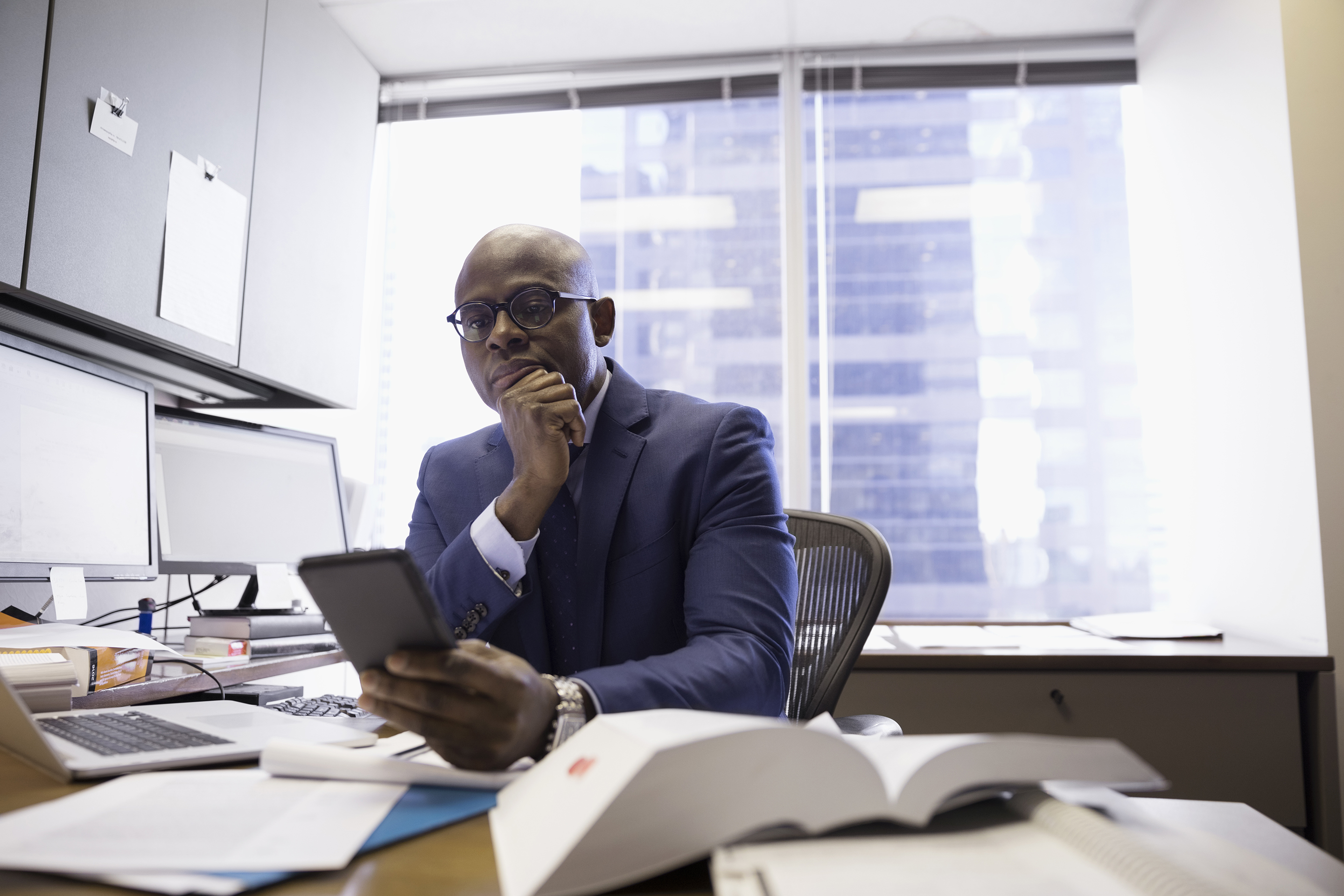 A professional man sits at his office desk looking at his phone.