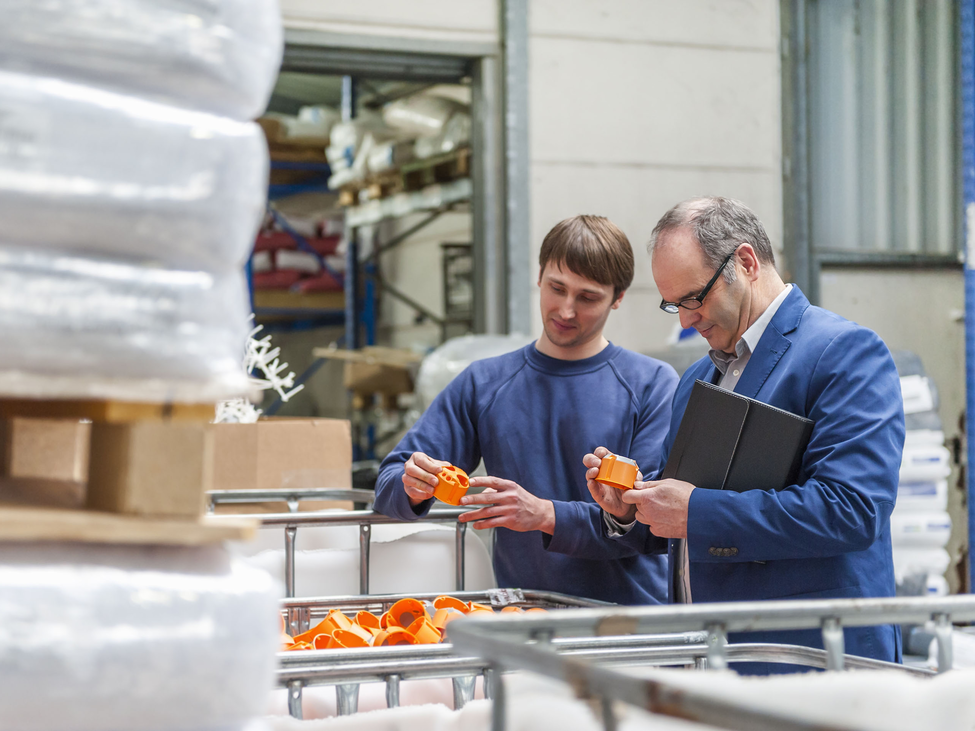 Two employees stand surrounded by stacks of plastic wrap on shelves while examining orange plastic components in a bin.