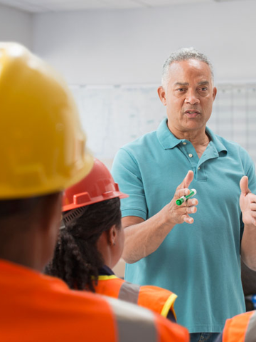 A manager speaks about safety and wellness to a group of employees wearing hard hats.