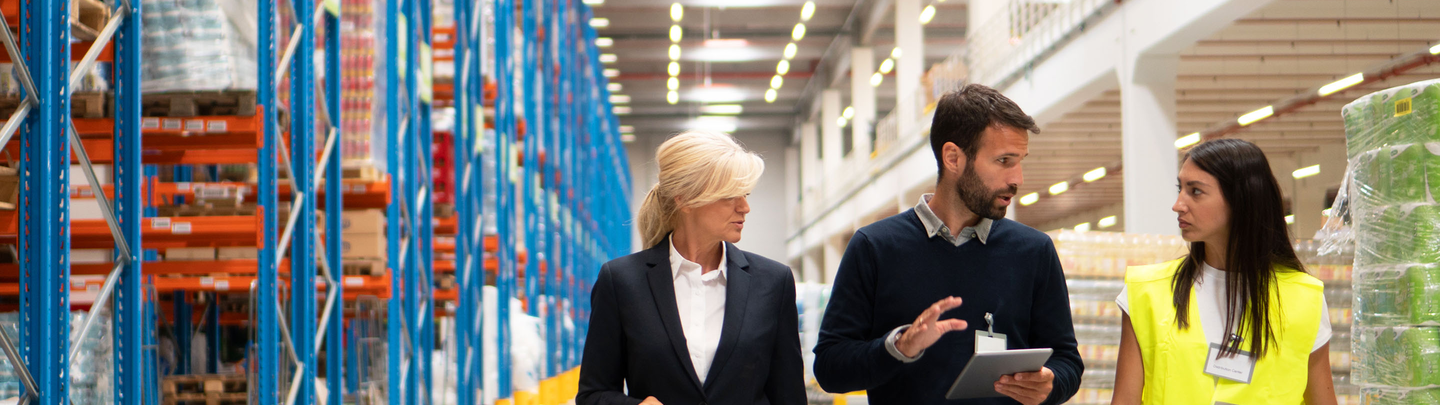 Insurance representative walks through warehouse with company employees, one in yellow reflective vest.