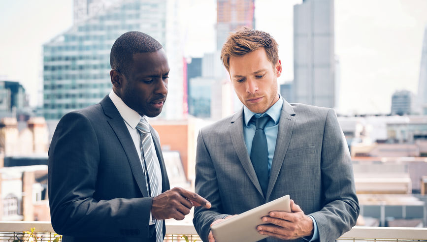 Businessman discussing project on digital tablet with his business partner, standing outdoor, city scape in the background.