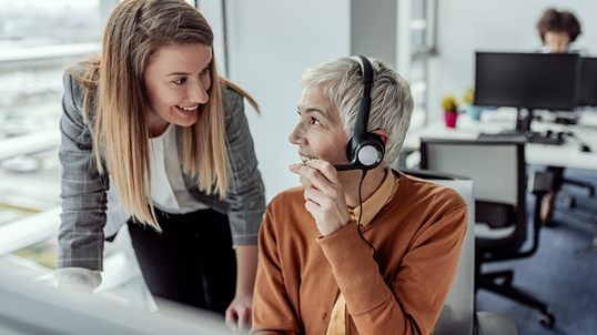 Two coworkers discussing something, one of the individuals is seated at a desk and is wearing a headset.