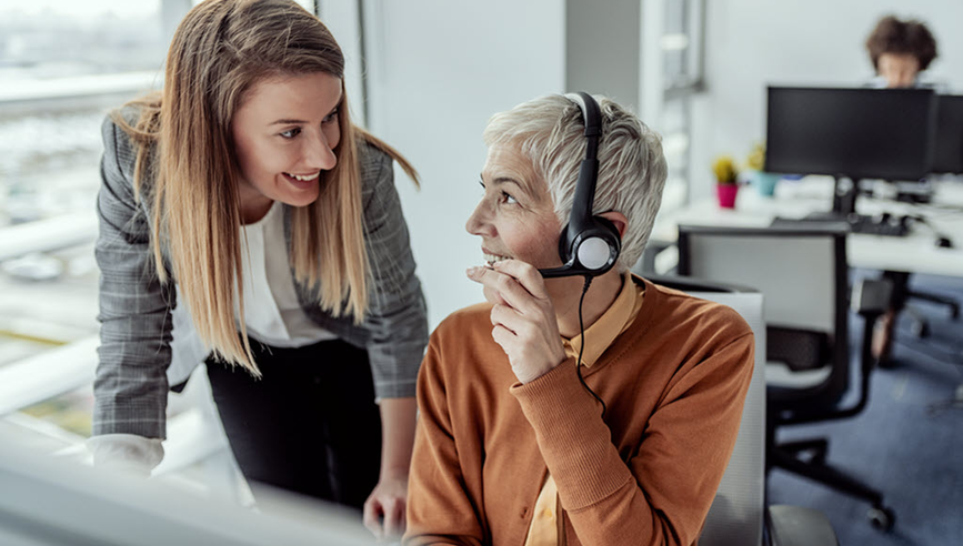 Two coworkers discussing something, one of the individuals is seated at a desk and is wearing a headset.