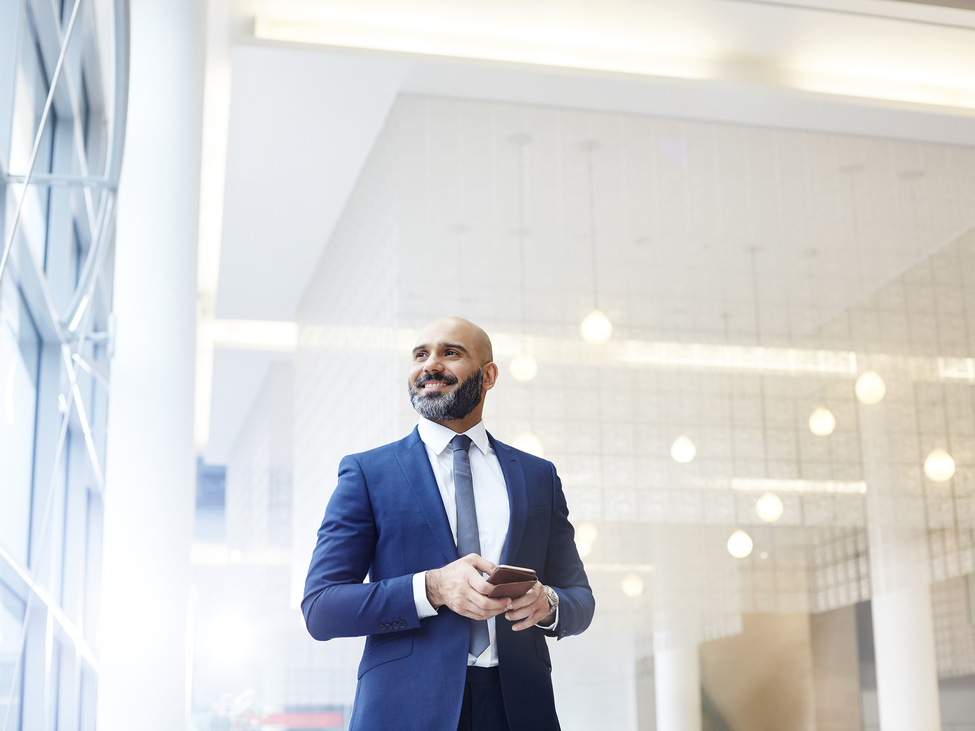 A finance professional holds a smartphone while looking toward a wall of windows in a modern office building.