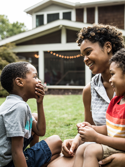 A mother and two elementary age school children giggle together while sitting in the front yard of their house.