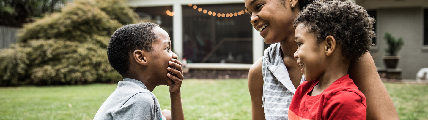 A mother and two elementary age school children giggle together while sitting in the front yard of their house.