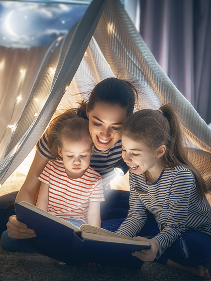 A mother reads to her children by flashlight while in a blanket fort.