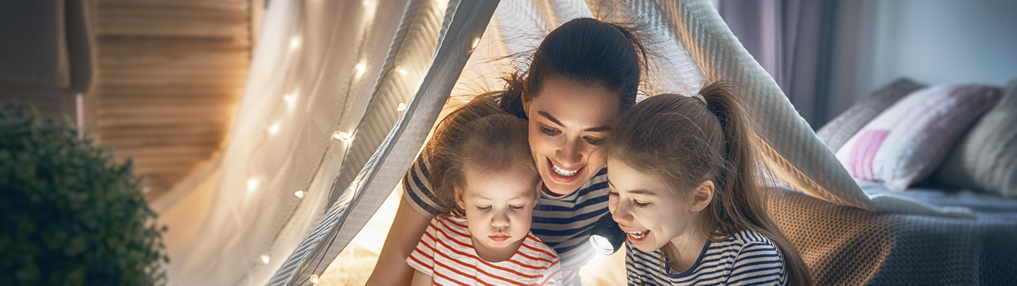 A mother reads to her children by flashlight while in a blanket fort.