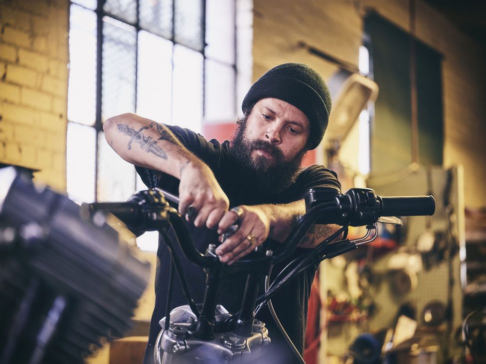 Man with beanie had and tattoos fixing a motorcycle in a shop.