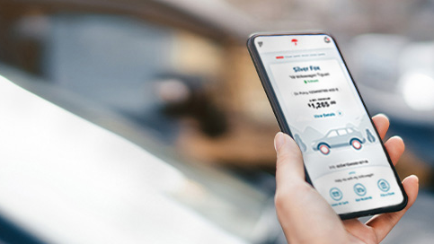 Woman holding mobile phone displaying MyTravelers app, car in background.