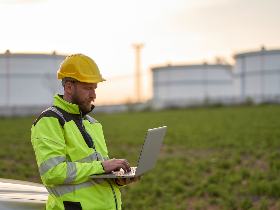 An energy technician in a hardhat and high-visibility safety jacket standing outside working on a laptop.