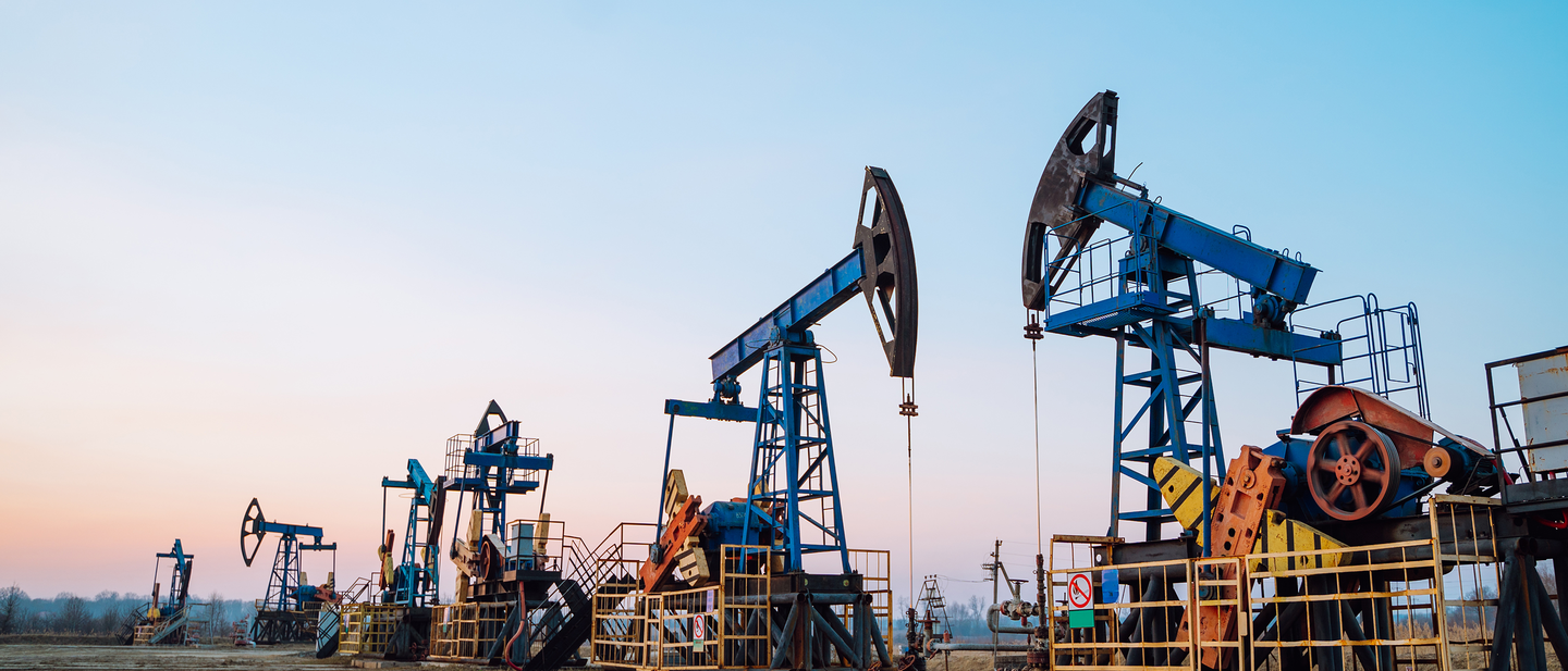 Pumpjacks line up on dry flatlands against a pastel-colored big sky.