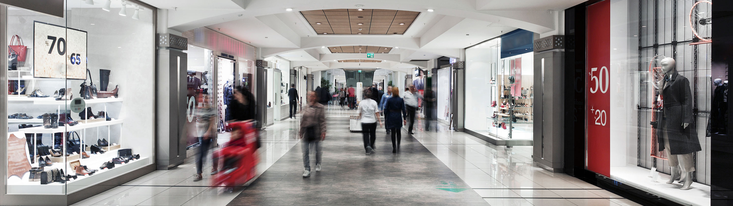 People walk inside a mall hallway with large chain retail stores on both sides.