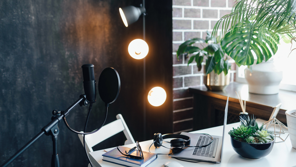 Podcast streaming at home. Audio studio with laptop, microphone with pop filter and headphones on white table against black wall with warm lights. Blogger concept