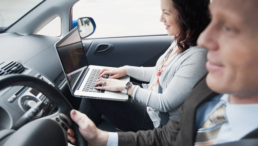 Professionals in car and passenger typing on laptop.