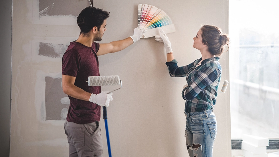 A man and woman looking at a color fan deck next to their house interior wall.
