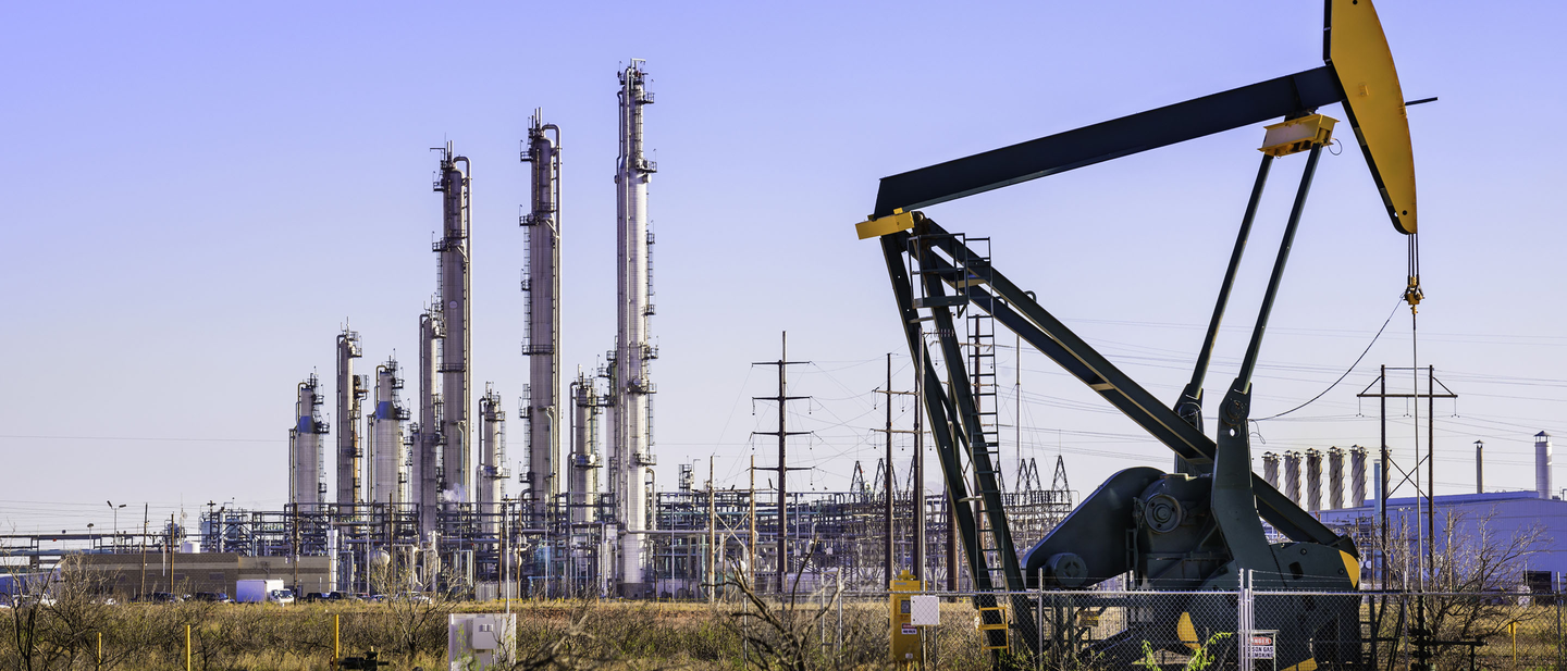 A pumpjack in the foreground with an oil refinery plant in the background.