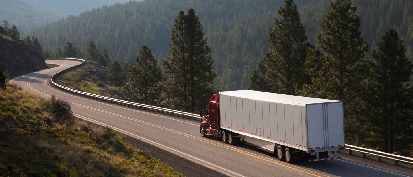 Red and white truck driving away on a remote highway surrounded by tall trees.