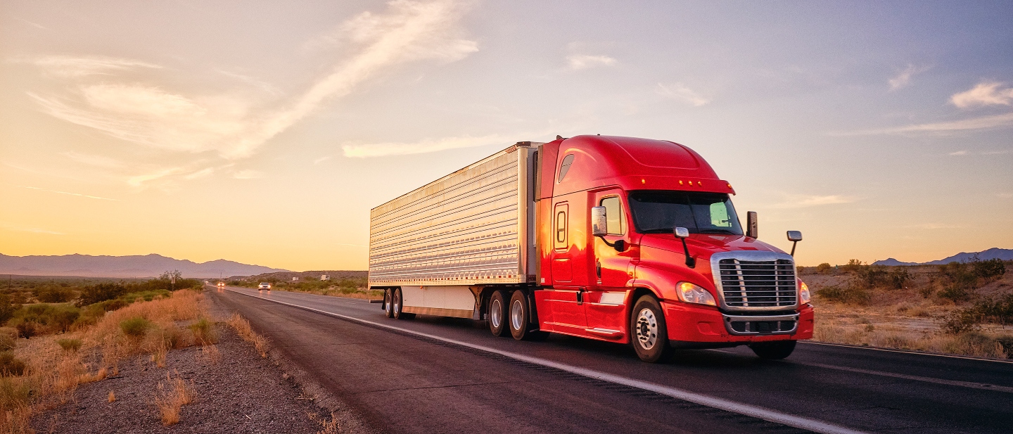 Red semitruck hauling freight on a rural highway under an evening sky.