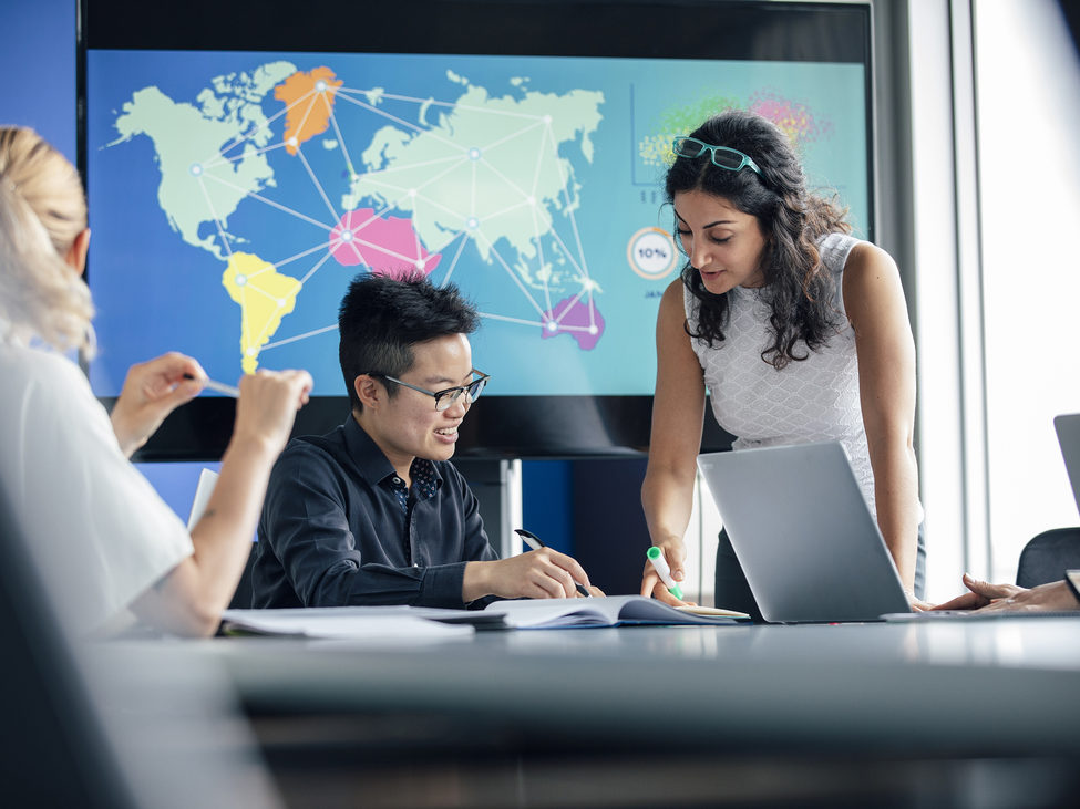 In a conference room, an international group of businesspeople discuss a report in front of a large map of the world showing business data.