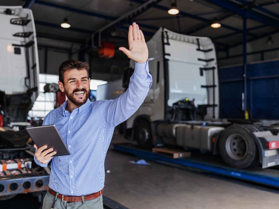 Smiling male shipping firm manager holding a digital tablet while waving from a warehouse of trucks.