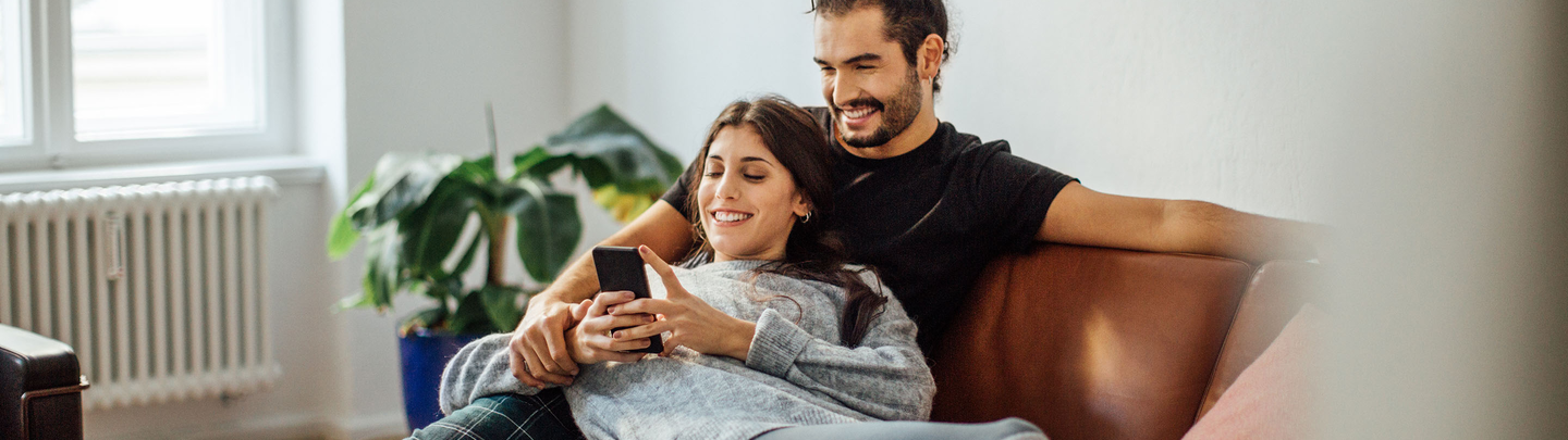 Happy young couple looking at a mobile phone while relaxing on sofa.