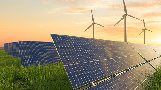 Solar panels in front of wind turbines in a field, with the sun setting in the background.