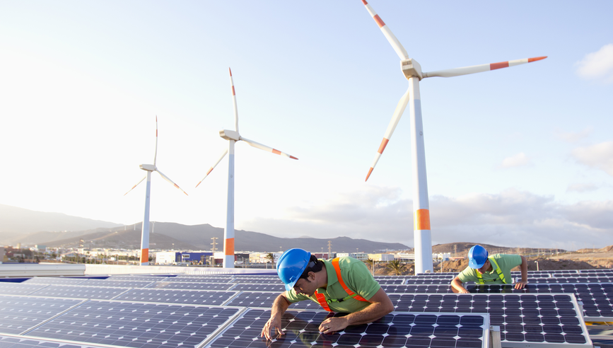 Image of workers inspecting solar panels at a renewable energy plan. In the background you can see Wind Turbines.