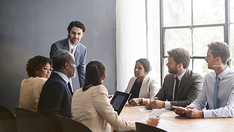 group of people sitting at a meeting table with a person talking and standing at end of table