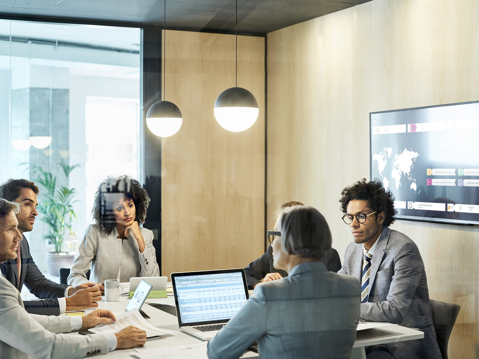 A consultant leads a presentation and discussion with a small group of business leaders and stakeholders in a glass-walled conference room.