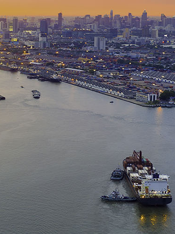 Wide shot of a container ship in a river along a busy shipping port as the sun sets behind a dense city skyline.