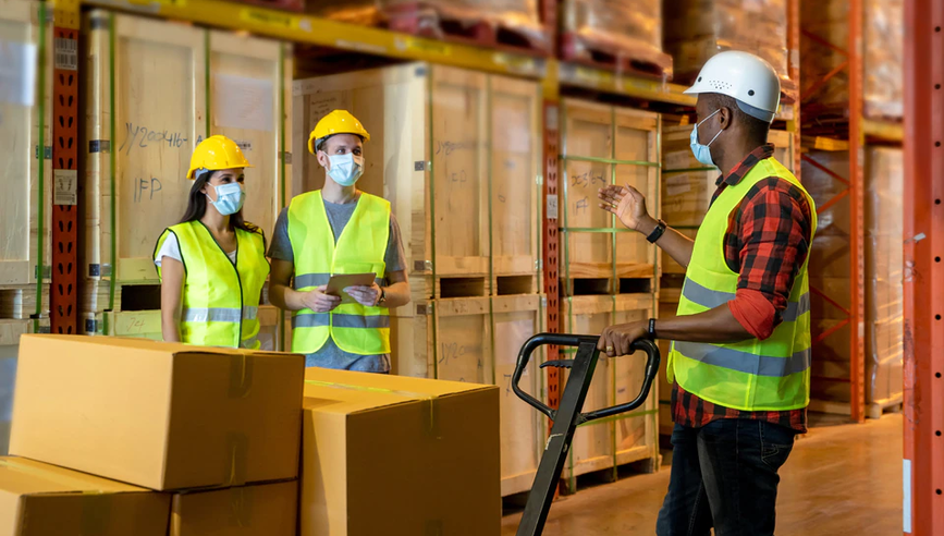 Three workers in a warehouse talking to each other with safety vests, hard hats, and face masks on.
