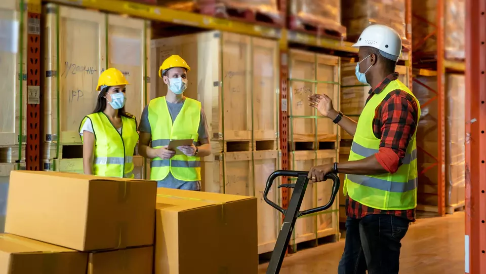 Three workers in a warehouse talking to each other with safety vests, hard hats and face masks on.