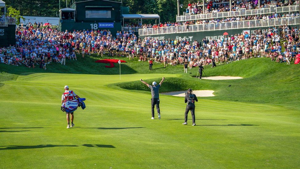 Travelers Championship golf tournament. Golfer with hand raised on field with crowd watching.