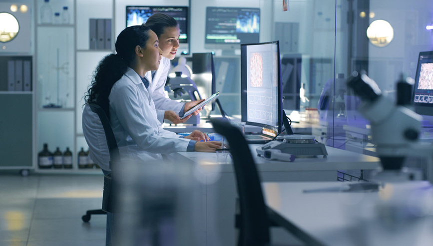 Two women working on a large desktop and tablet in a science lab.