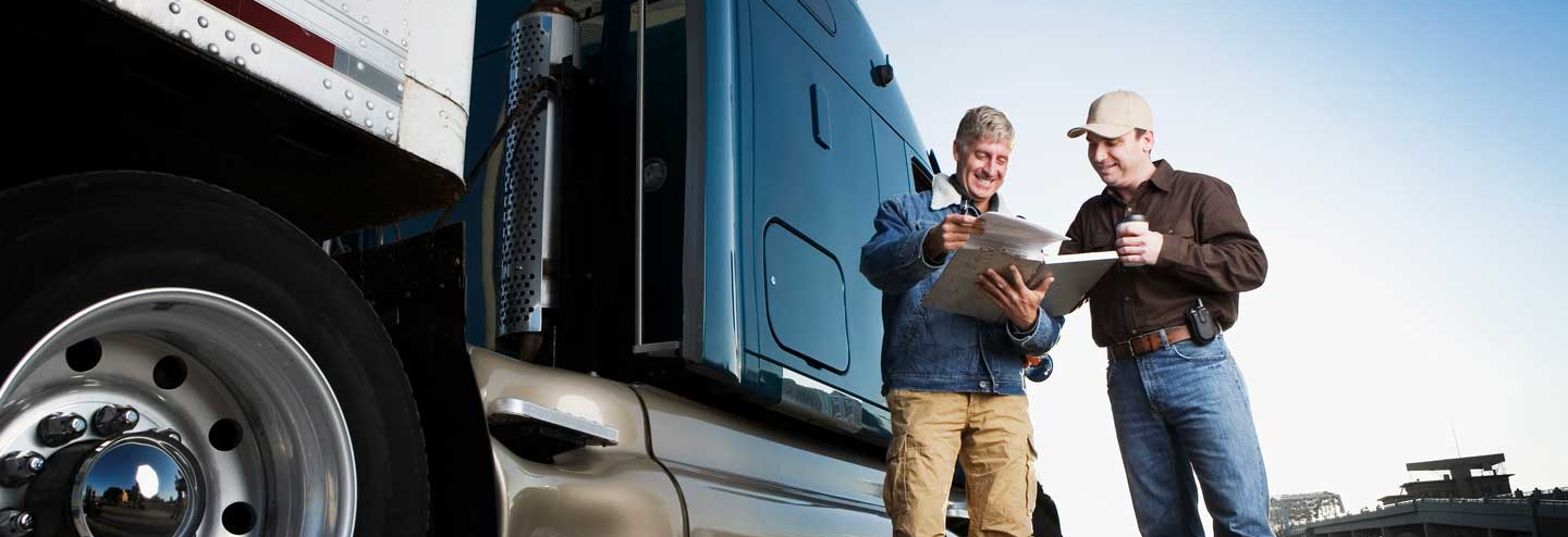 Two male drivers smiling and looking at paperwork while standing in front of a blue and white truck.