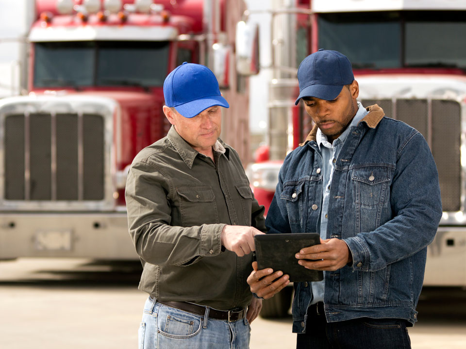 Two male truck drivers wearing blue hats have a discussion while looking at a tablet. 
