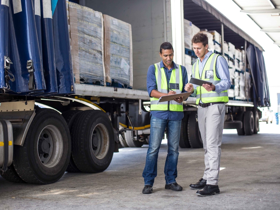 Two male workers standing behind a loaded truck at a large warehouse, looking at a clipboard together.