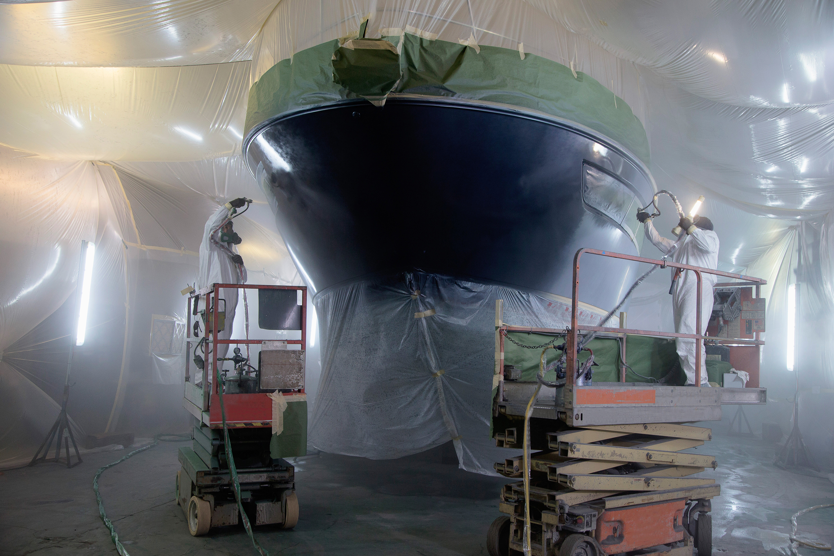 Two men paint boat in warehouse.